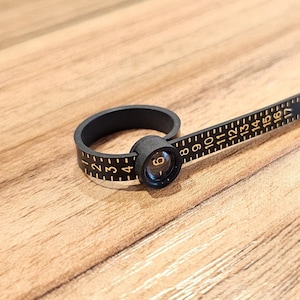 May include: Black ring sizer with a flexible ruler marked with numbers from 1 to 17. The ruler is attached to a black ring that can be adjusted to fit different finger sizes.