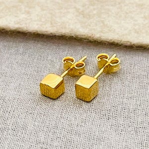 May include: A pair of gold-colored cube stud earrings. Each earring features a small, square-shaped charm suspended from a post. The earrings are displayed on a textured, light-colored surface.
