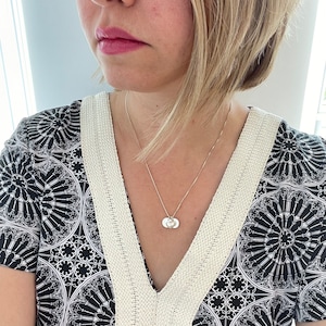 Peut inclure: Un collier en argent avec une chaîne délicate et un pendentif composé de deux petits disques ronds. Le collier est porté par une personne aux cheveux blonds courts et portant une chemise à motifs noir et blanc.