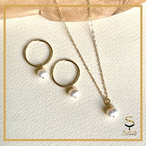 May include: A gold-coloured jewellery set comprising a necklace and hoop earrings, each with a single white pearl. The necklace has a fine chain, and the earrings are circular. The jewellery is presented on a light-coloured surface.