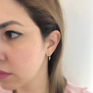 May include: A close-up of a person's ear with a gold lightning bolt dangle earring. The earring is small and delicate, and the lightning bolt is a simple design.