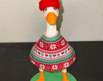 May include: A whimsical goose figurine wearing a red knit hat and a festive sweater. The sweater features a pattern of white snowflakes and red hearts on a green background. The goose has orange feet and a green base.