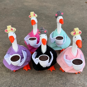 May include: Six white goose figurines wearing colorful robes and hats, each holding a white cup with brown liquid. The geese are arranged in a semi-circle on a gray surface.