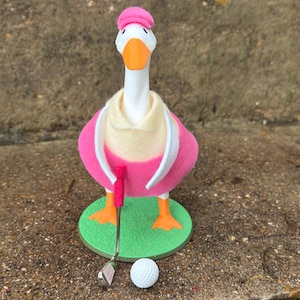 May include: A white goose figurine wearing a pink hat, a cream-colored collared shirt, and a pink vest. The goose stands on a green circular base, holding a miniature golf club with a pink grip next to a small white dimpled golf ball.