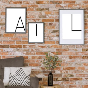 Könnte beinhalten: Ein grauer Sessel mit einem weißen und schwarzen geometrischen Musterkissen steht vor einer Backsteinwand. Drei gerahmte Drucke mit den Buchstaben "A", "T" und "L" hängen an der Wand. Ein kleiner Holz-Beistelltisch mit schwarzen Metallbeinen steht neben dem Sessel und trägt eine klare Glasvase mit einer grünen, belaubten Pflanze.