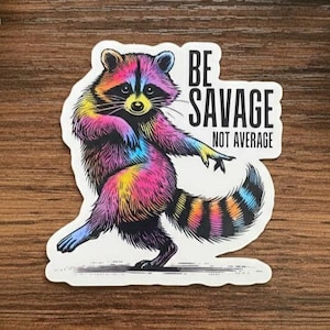 Peut inclure: Autocollant représentant un raton laveur coloré avec une fourrure et une queue arc-en-ciel. Le raton laveur est en position de danse avec le texte "BE SAVAGE NOT AVERAGE" dans un encadré blanc. Fond en bois et plantes vertes.