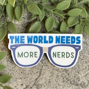 May include: A white sticker with blue and green text that reads "The World Needs More Nerds". The text is displayed on a pair of blue-rimmed glasses.