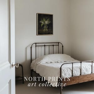 May include: A white bed with a metal frame and a white linen duvet cover. A framed print of a bouquet of white flowers in a glass vase hangs on the wall above the bed. The text "NORTH PRINTS art collective" is printed on the floor in front of the bed.