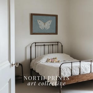 May include: A white butterfly print in a wooden frame hangs above a white bed with a metal frame and a white bedspread. A teddy bear lies on the bed. The text "NORTH PRINTS art collective" is at the bottom of the image.