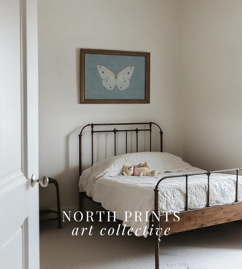 May include: A white butterfly print in a wooden frame hangs above a white bed with a metal frame and a white bedspread. A teddy bear lies on the bed. The text "NORTH PRINTS art collective" is at the bottom of the image.