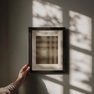 May include: A framed print featuring a brown and beige plaid pattern. The artwork is displayed in a dark wooden frame and held by a person's hand. The wall behind the frame is a neutral gray color, with shadows cast across it.