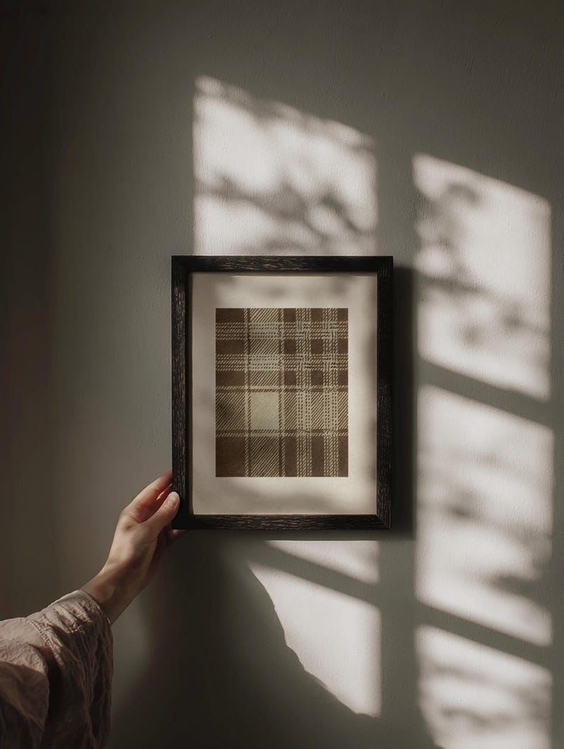 May include: A framed print featuring a brown and beige plaid pattern. The artwork is displayed in a dark wooden frame and held by a person's hand. The wall behind the frame is a neutral gray color, with shadows cast across it.
