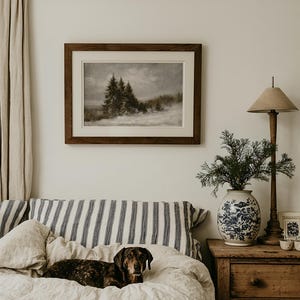 Puede incluir: Una impresi&oacute;n de paisaje enmarcada que muestra una escena forestal oscura y melanc&oacute;lica con suelo cubierto de nieve, presentada en un marco de madera oscura con paspart&uacute; blanco. La obra cuelga sobre una cama con almohadas de rayas azules y blancas.