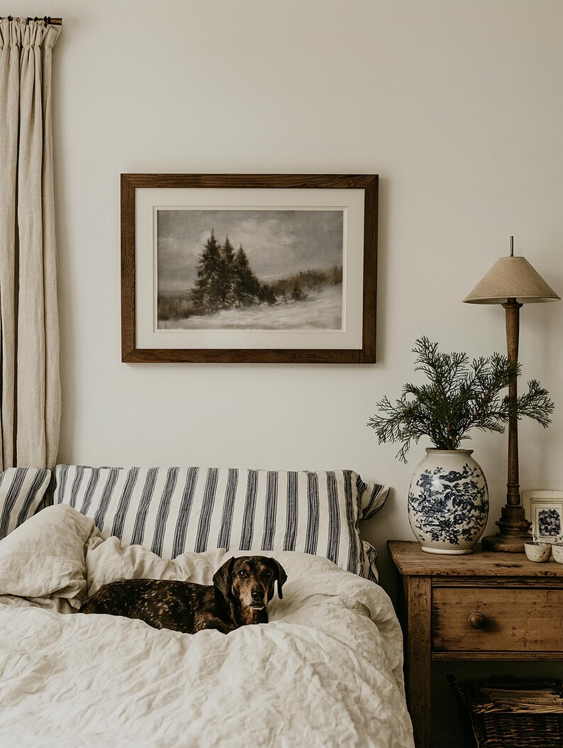 Puede incluir: Una impresi&oacute;n de paisaje enmarcada que muestra una escena forestal oscura y melanc&oacute;lica con suelo cubierto de nieve, presentada en un marco de madera oscura con paspart&uacute; blanco. La obra cuelga sobre una cama con almohadas de rayas azules y blancas.