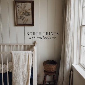 Op de afbeelding: Een ingelijste bloemenprint hangt aan een witte wand met panelen. Daaronder zijn een witte wieg met een cr&egrave;mekleurige deken en een kleine houten kruk met een gevlochten mand zichtbaar. De tekst "NORTH PRINTS art collective" staat onderaan.