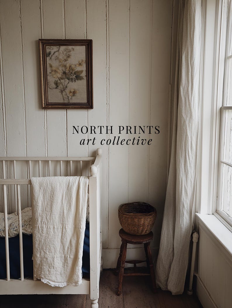 Op de afbeelding: Een ingelijste bloemenprint hangt aan een witte wand met panelen. Daaronder zijn een witte wieg met een cr&egrave;mekleurige deken en een kleine houten kruk met een gevlochten mand zichtbaar. De tekst "NORTH PRINTS art collective" staat onderaan.