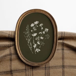 May include: An oval framed botanical print featuring white floral illustrations on a dark green background. The frame is gold-colored, and the print is displayed against a brown plaid fabric.