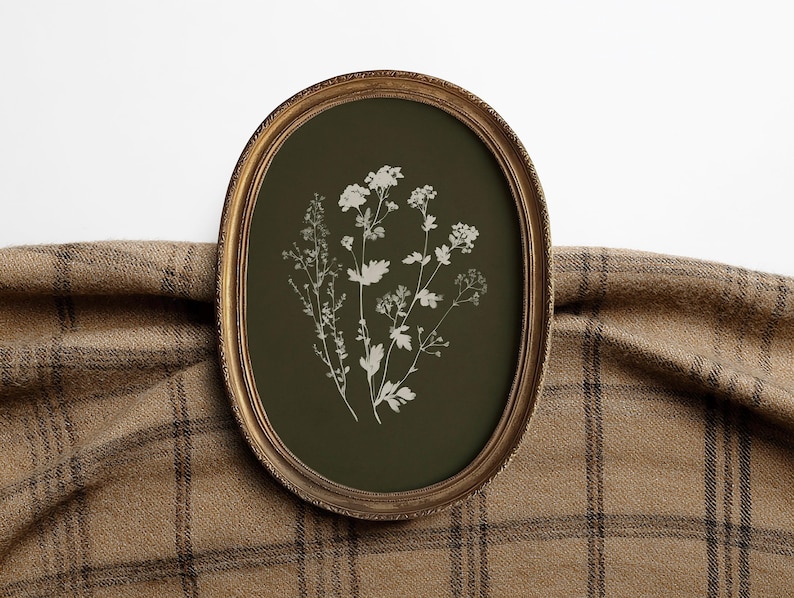 May include: An oval framed botanical print featuring white floral illustrations on a dark green background. The frame is gold-colored, and the print is displayed against a brown plaid fabric.