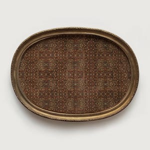 May include: An oval serving tray with a decorative gold-coloured frame. The tray's interior features a detailed pattern in shades of brown, red, and beige. The frame has an ornate, textured design. The tray is set against a plain white background.