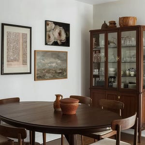 May include: A dark wood oval dining table with six chairs. The table has a smooth, polished surface. The chairs are upholstered in a light beige fabric. There is a wooden cabinet with glass doors in the background. The cabinet is filled with dishes and glassware. There are several framed pictures on the wall behind the table.