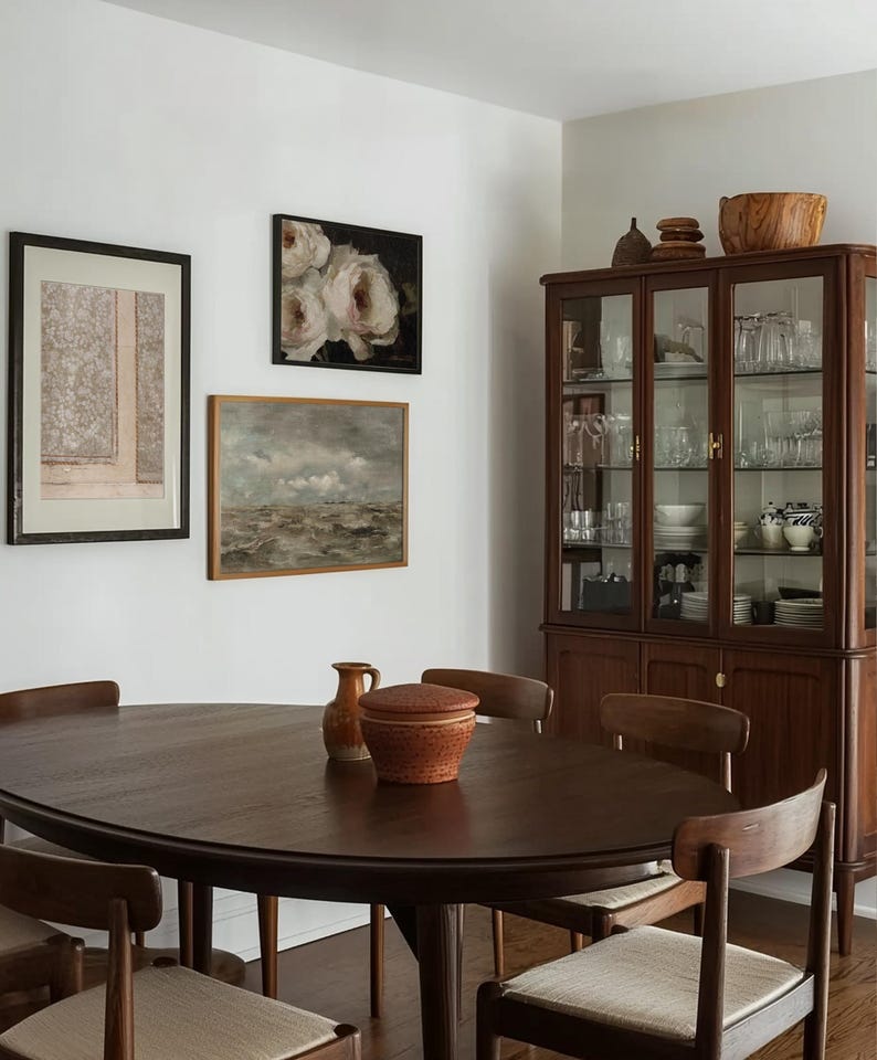 May include: A dark wood oval dining table with six chairs. The table has a smooth, polished surface. The chairs are upholstered in a light beige fabric. There is a wooden cabinet with glass doors in the background. The cabinet is filled with dishes and glassware. There are several framed pictures on the wall behind the table.