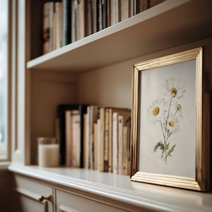 Op de afbeelding: Een gouden lijst met een bloemenprint van witte madeliefjes met gele centra en groene stelen. De lijst staat op een plank met boeken, een kaars en een raam. De print voegt een natuurlijk element toe aan de kamer.