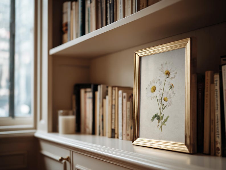 Op de afbeelding: Een gouden lijst met een bloemenprint van witte madeliefjes met gele centra en groene stelen. De lijst staat op een plank met boeken, een kaars en een raam. De print voegt een natuurlijk element toe aan de kamer.