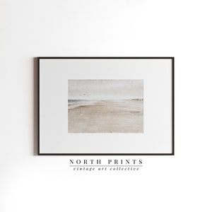 May include: A framed print of a watercolour painting depicting a beach scene with a sandy shore, a calm ocean, and a few birds flying in the sky. The print is framed in a black frame and has the text "NORTH PRINTS vintage art collective" at the bottom.