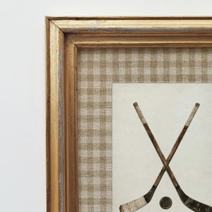 May include: Framed artwork featuring two crossed hockey sticks and a puck. The artwork has a gold-colored frame with a distressed finish. The background is a tan and white checkered pattern, with a cream-colored center.