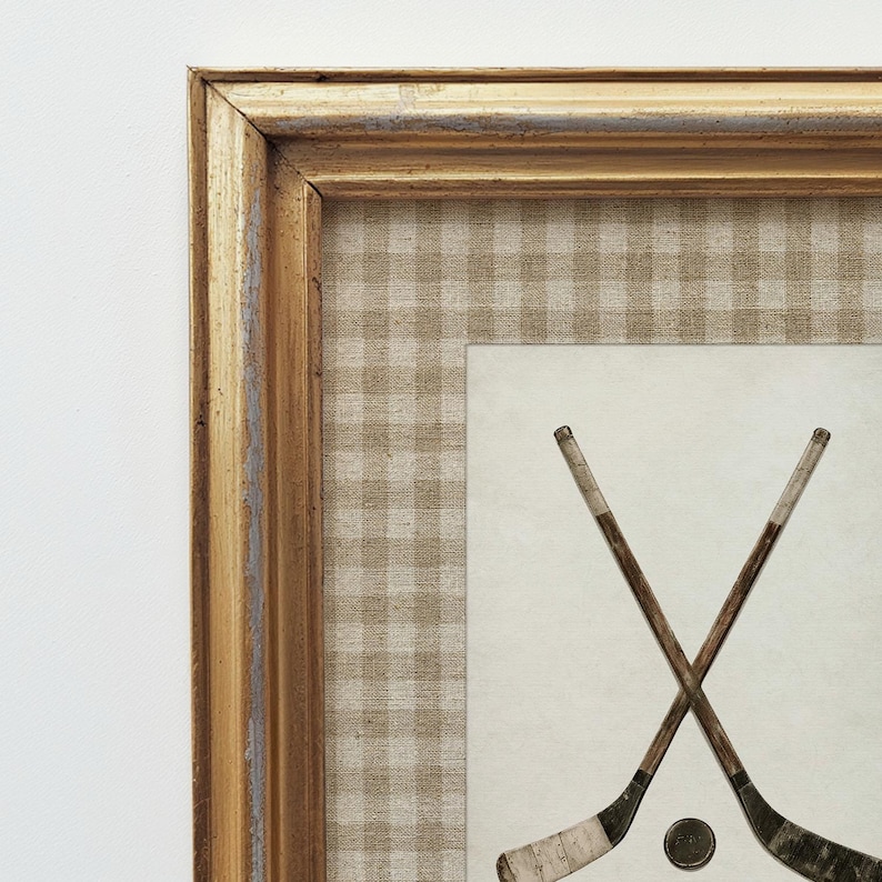 May include: Framed artwork featuring two crossed hockey sticks and a puck. The artwork has a gold-colored frame with a distressed finish. The background is a tan and white checkered pattern, with a cream-colored center.
