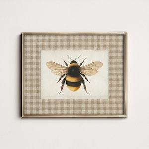 May include: A framed print featuring a detailed watercolor illustration of a bumblebee. The bee is depicted with black and yellow stripes, brown wings, and delicate legs. The print is set within a gold-colored frame and a tan and white checkered mat.