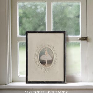May include: A framed black and white illustration of a ballerina in a white tutu, standing on one leg with her arms outstretched. The illustration is surrounded by a decorative white frame with a floral design. The frame is hanging on a white wall.