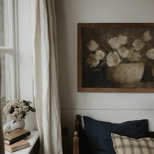 May include: A framed painting of white flowers in a vase hangs above a bed with navy blue and plaid pillows. A window with a white curtain is on the left. A vase of flowers and books sit on the windowsill.