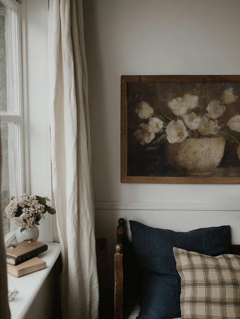 May include: A framed painting of white flowers in a vase hangs above a bed with navy blue and plaid pillows. A window with a white curtain is on the left. A vase of flowers and books sit on the windowsill.