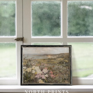 Peut inclure: Une peinture encadr&eacute;e repr&eacute;sentant un paysage c&ocirc;tier avec un champ de fleurs roses et blanches au premier plan. La peinture est dans un cadre noir et est pos&eacute;e sur un rebord de fen&ecirc;tre blanc. Le texte "NORTH PRINTS art collective" est sous la peinture.