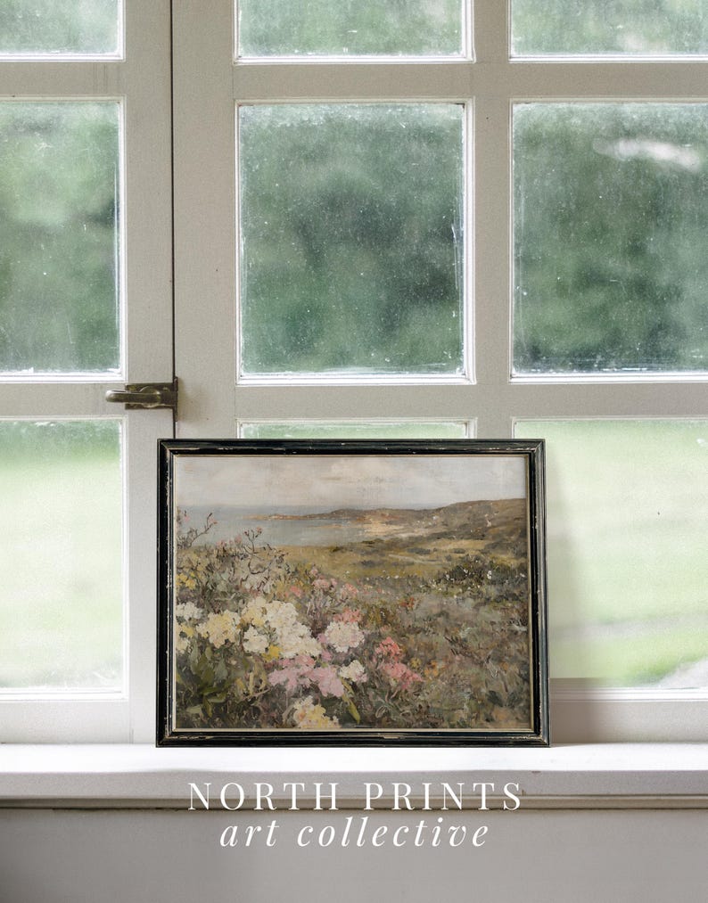 Peut inclure: Une peinture encadr&eacute;e repr&eacute;sentant un paysage c&ocirc;tier avec un champ de fleurs roses et blanches au premier plan. La peinture est dans un cadre noir et est pos&eacute;e sur un rebord de fen&ecirc;tre blanc. Le texte "NORTH PRINTS art collective" est sous la peinture.