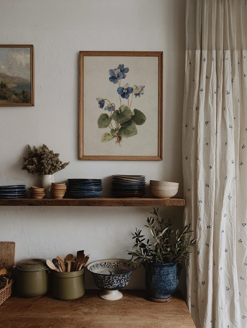 K&ouml;nnte beinhalten: Ein gerahmter botanischer Druck mit blauen Veilchen h&auml;ngt &uuml;ber einem Holzregal mit Tellern und Schalen. Eine rustikale K&uuml;chenszene mit olivgr&uuml;nen Beh&auml;ltern, Holzutensilien und einer blau-wei&szlig; gemusterten Schale.