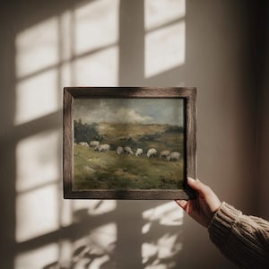 K&ouml;nnte beinhalten: Gerahmtes Landschaftsgem&auml;lde mit Schafen, die auf einer gr&uuml;nen Wiese unter einem bew&ouml;lkten Himmel grasen. Das Kunstwerk wird in einem dunklen Holzrahmen pr&auml;sentiert und erinnert an eine Vintage-&Auml;sthetik.