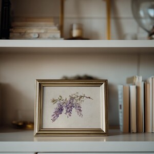 May include: A framed botanical print featuring a watercolor illustration of purple wisteria blossoms. The gold-toned frame sits on a white shelf, surrounded by books. The artwork adds a touch of nature-inspired decor.