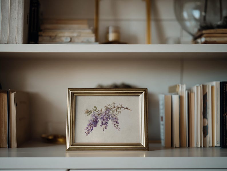 May include: A framed botanical print featuring a watercolor illustration of purple wisteria blossoms. The gold-toned frame sits on a white shelf, surrounded by books. The artwork adds a touch of nature-inspired decor.