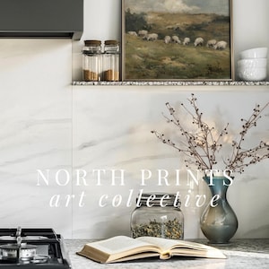 K&ouml;nnte beinhalten: K&uuml;chenszene mit Landschaftsbild von Schafen. Ein Glas, eine Vase mit Zweigen und ein offenes Buch liegen auf der Arbeitsplatte. Der Text "NORTH PRINTS art collective" ist zu sehen. Neutrale Farbgebung.