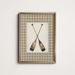 Puede incluir: Impresión enmarcada con una ilustración en acuarela de dos remos cruzados. Los remos de madera tienen detalles rojos y azules. El marco es dorado con un patrón de cuadros beige y blanco.