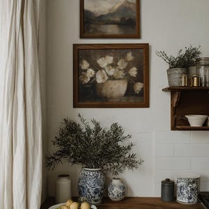 May include: A cozy kitchen scene featuring two framed landscape paintings and a floral still life. A wooden countertop displays a white bowl of lemons, blue and white vases, and a large vase of greenery. A white curtain hangs on the left.