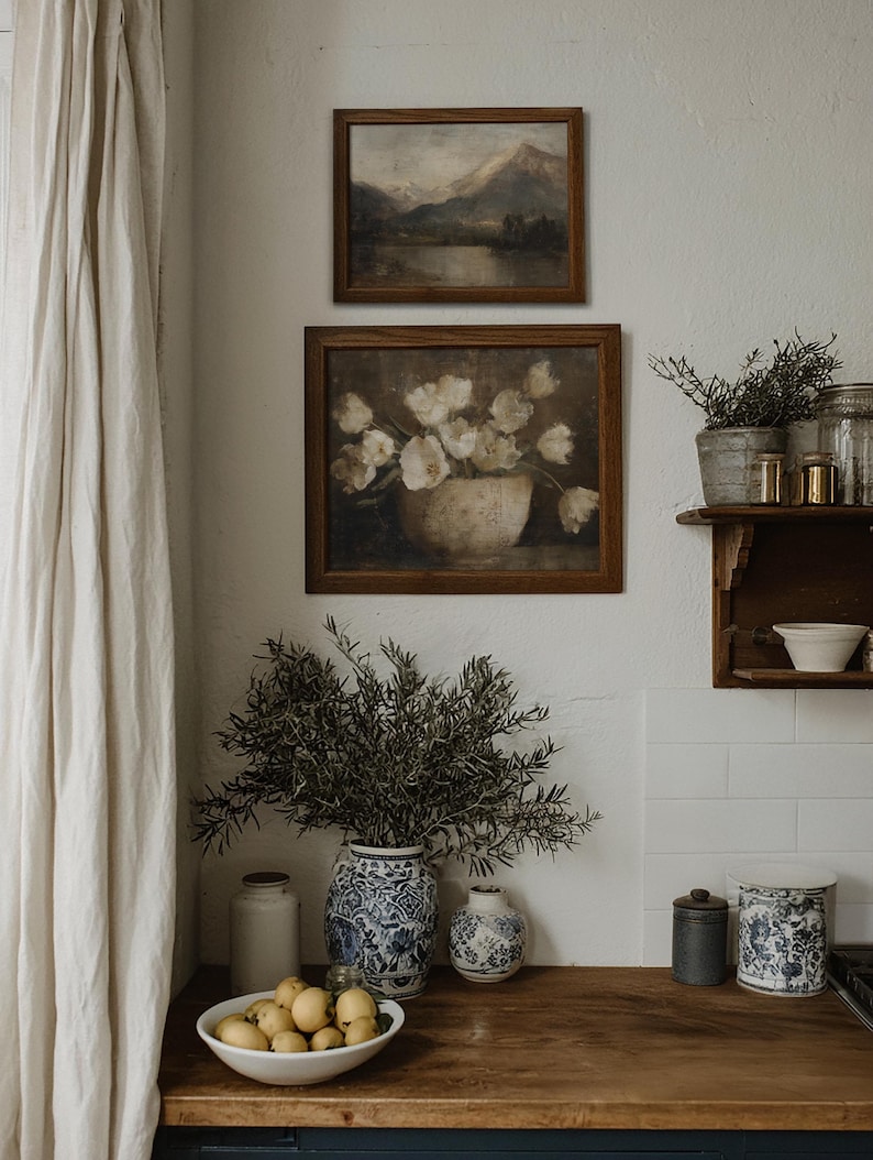 May include: A cozy kitchen scene featuring two framed landscape paintings and a floral still life. A wooden countertop displays a white bowl of lemons, blue and white vases, and a large vase of greenery. A white curtain hangs on the left.