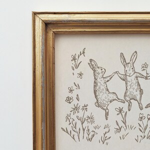 May include: A framed print of two rabbits dancing in a field of flowers. The rabbits are drawn in a simple, whimsical style, and the flowers are delicate and charming. The frame is a simple gold frame.