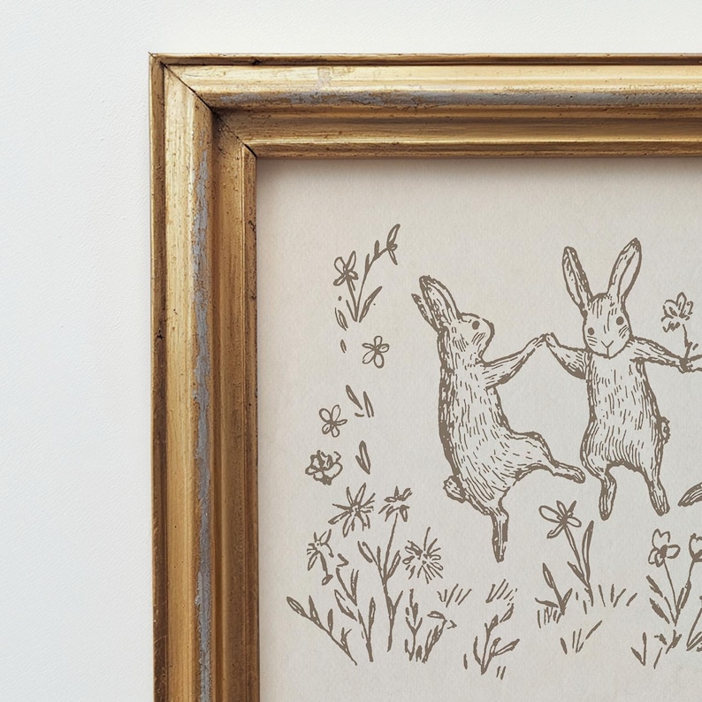 May include: A framed print of two rabbits dancing in a field of flowers. The rabbits are drawn in a simple, whimsical style, and the flowers are delicate and charming. The frame is a simple gold frame.
