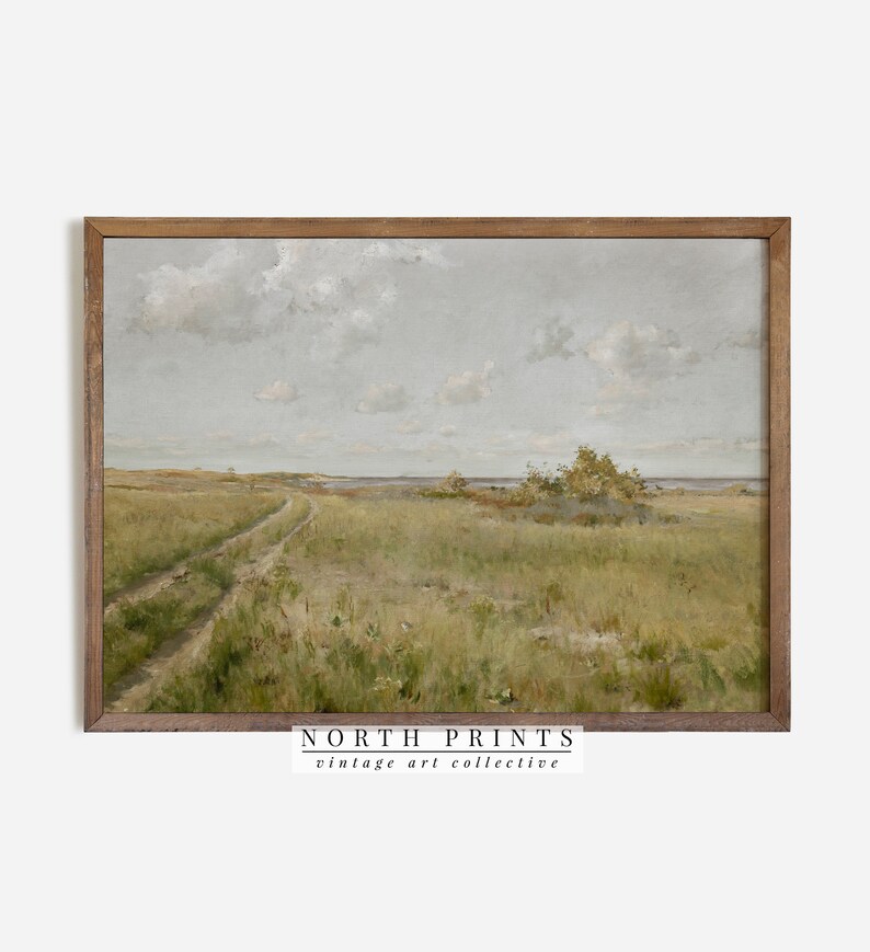 May include: A vintage landscape painting depicting a dirt road leading through a grassy field towards a body of water. The sky is a light blue with white clouds. The painting is framed in a brown wood frame.