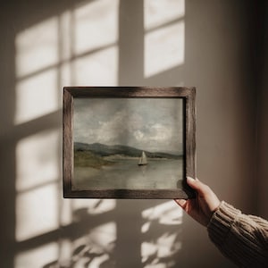 K&ouml;nnte beinhalten: Gerahmtes Landschaftsgem&auml;lde mit einem Segelboot auf einem See und Bergen im Hintergrund. Das Kunstwerk befindet sich in einem Holzrahmen. Die Farbpalette umfasst Graut&ouml;ne, Gr&uuml;n und Braun.