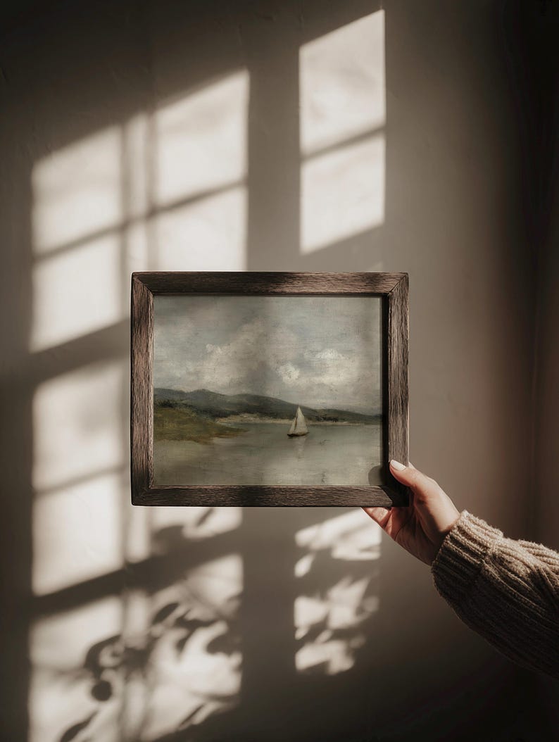 K&ouml;nnte beinhalten: Gerahmtes Landschaftsgem&auml;lde mit einem Segelboot auf einem See und Bergen im Hintergrund. Das Kunstwerk befindet sich in einem Holzrahmen. Die Farbpalette umfasst Graut&ouml;ne, Gr&uuml;n und Braun.