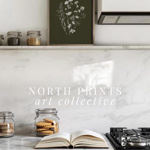 May include: A framed botanical print with white floral illustrations on a dark green background hangs above a countertop. Jars of ingredients and an open book with cookies are on the counter. The text "NORTH PRINTS art collective" is visible.